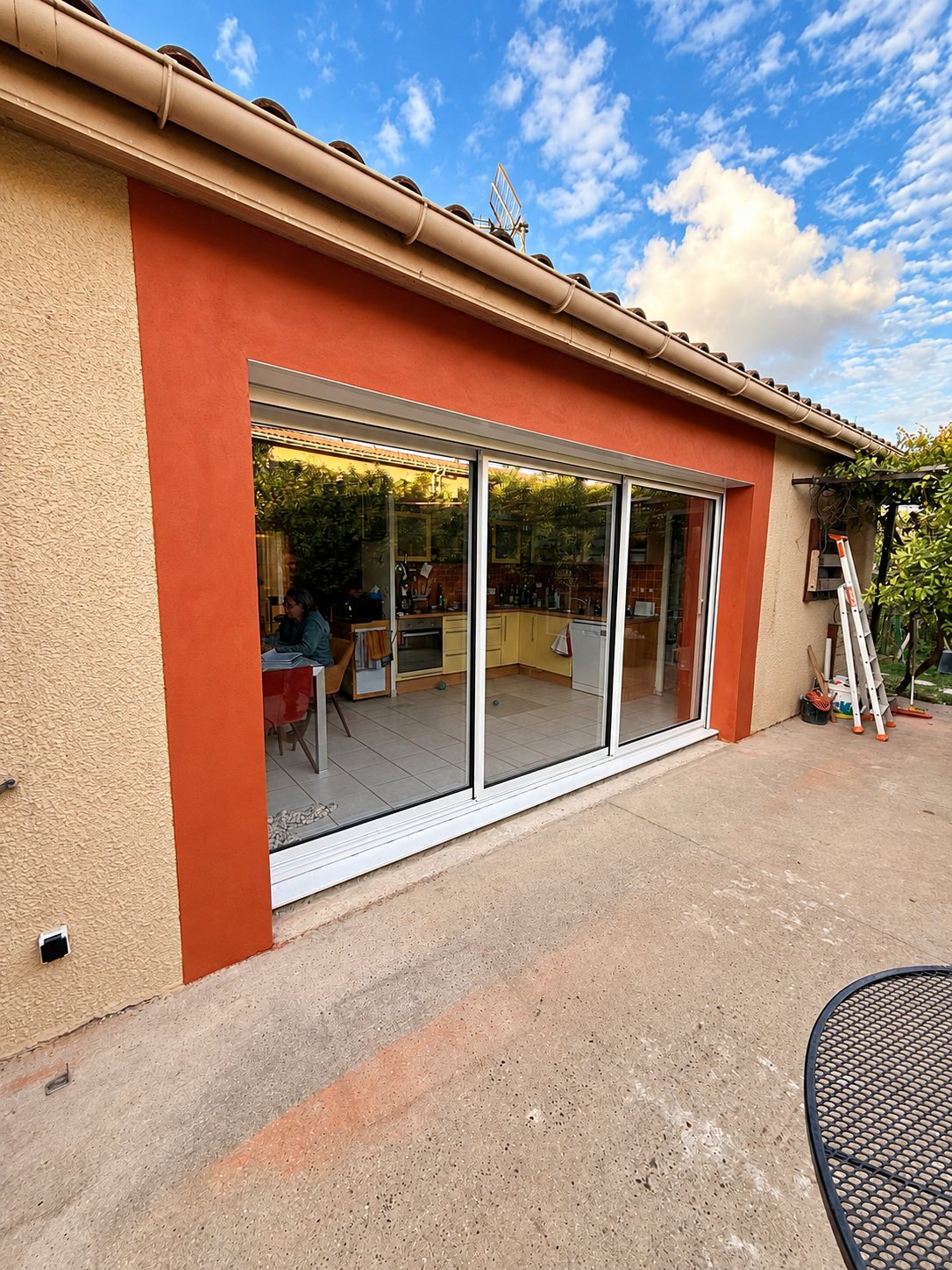 Une grande baie vitrée coulissante à cadre blanc encadrée d'un enduit orange sur la façade d'une maison, avec une terrasse en béton au premier plan.