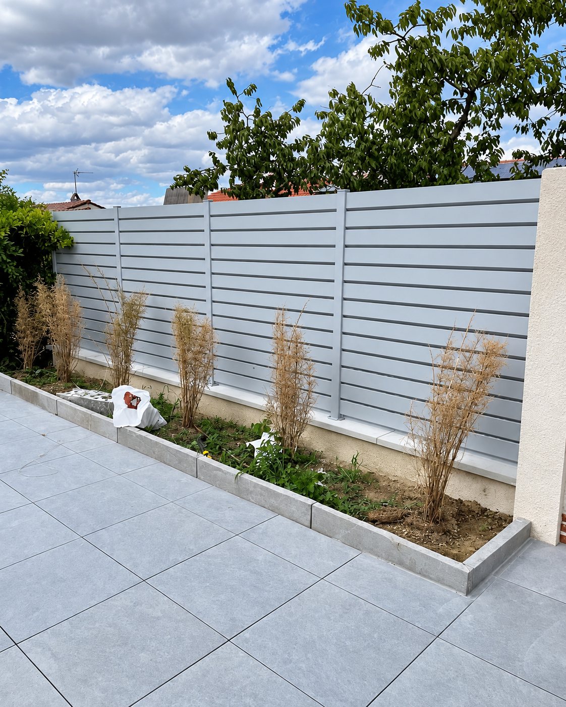 Une clôture en aluminium gris à lames horizontales borde une terrasse en grandes dalles grises, avec un massif de jeunes graminées au premier plan.