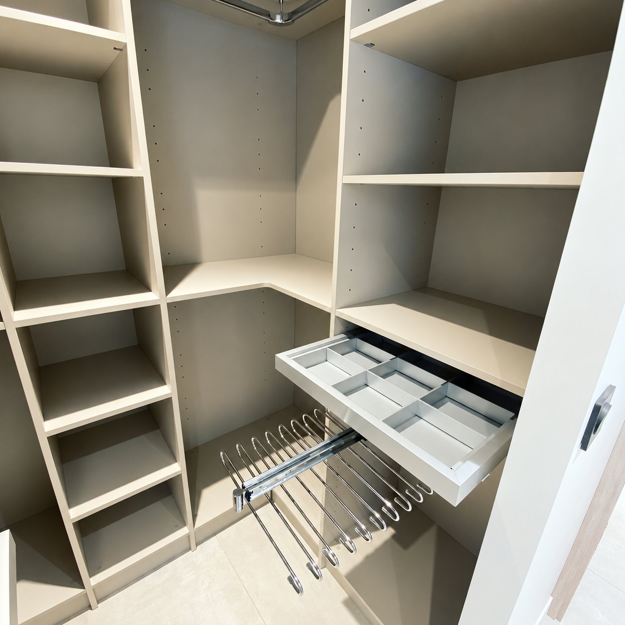 Intérieur d'un dressing d'angle beige avec étagères, un tiroir compartimenté sorti et un porte-pantalons chromé coulissant.