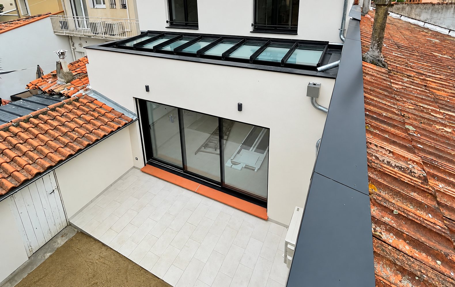 Vue en hauteur d'une maison rénovée avec verrière sur le toit, grande baie vitrée coulissante noire et petite terrasse carrelée entourée de toits en tuiles.