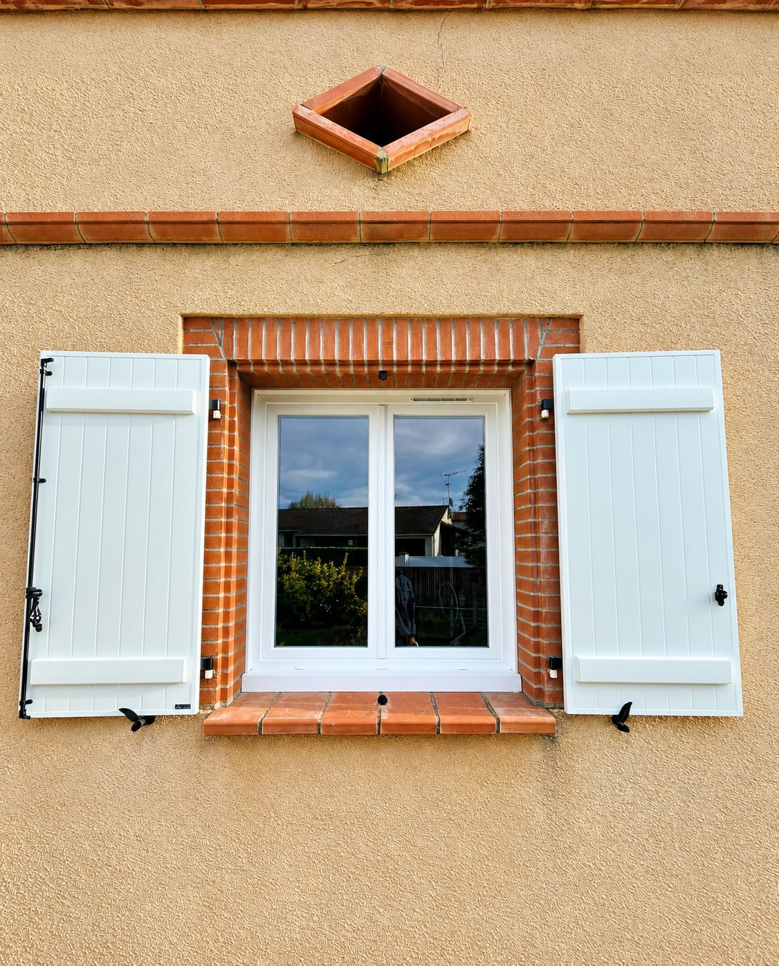 Fenêtre en PVC blanc encadrée de briques rouges et de deux volets blancs ouverts sur une façade en crépi beige.