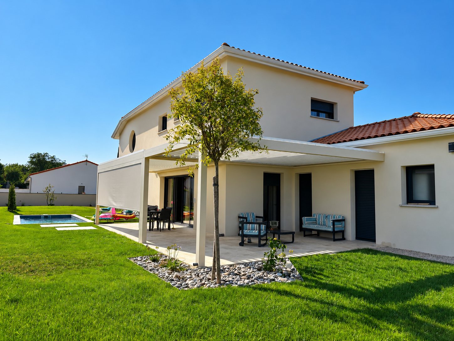 Maison crépie à étage avec une pergola à lames orientables au-dessus d'une terrasse meublée, un jardin engazonné et une petite piscine.