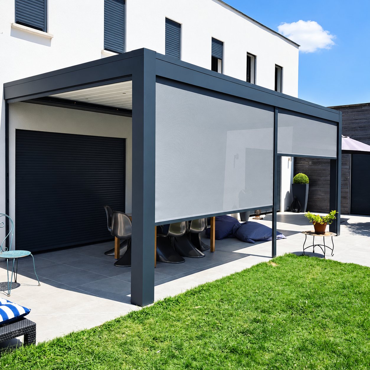 Une pergola en aluminium gris anthracite équipée de stores verticaux en toile couvre une terrasse carrelée devant une maison blanche moderne, avec une pelouse au premier plan.
