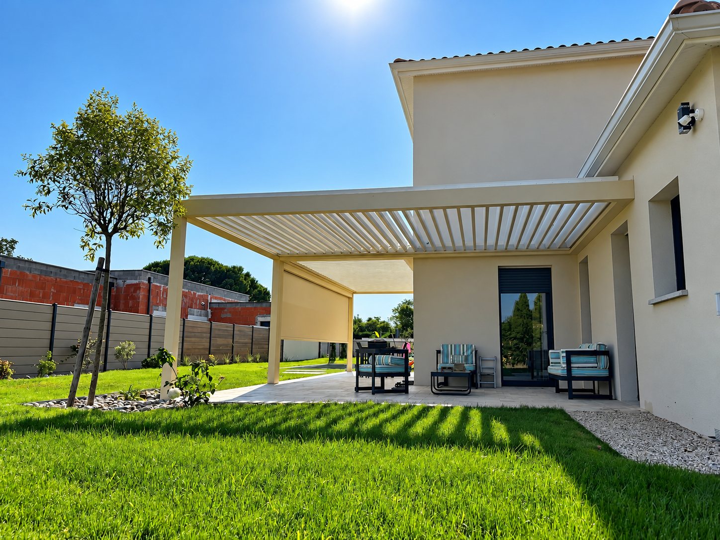 Une pergola bioclimatique beige à lames orientables couvre une terrasse meublée de fauteuils, devant une maison récente et une pelouse verte ensoleillée.