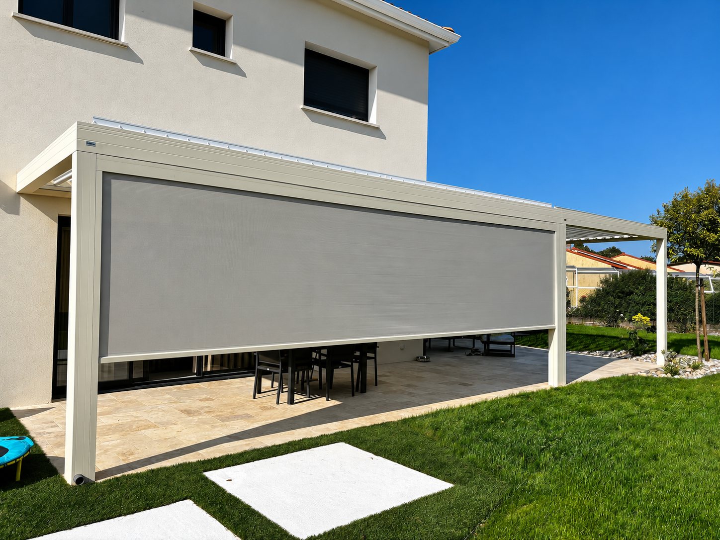 Une pergola en aluminium beige équipée d'un grand store vertical gris abrite une terrasse en dalles de pierre devant une maison avec jardin.