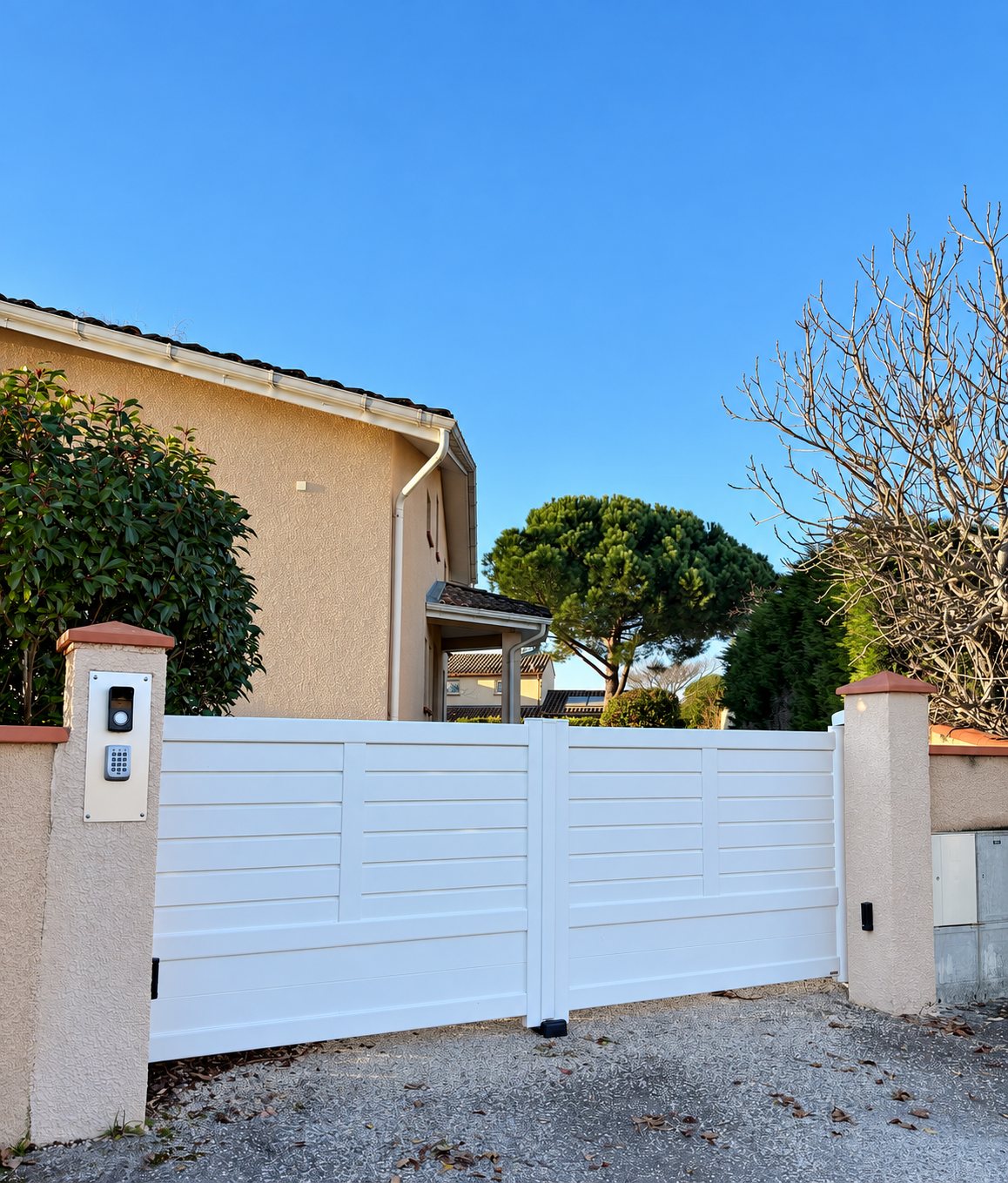 Un portail battant blanc à lames horizontales installé entre deux piliers crépis, dont l'un porte un interphone et un clavier à code, devant une maison au crépi beige.