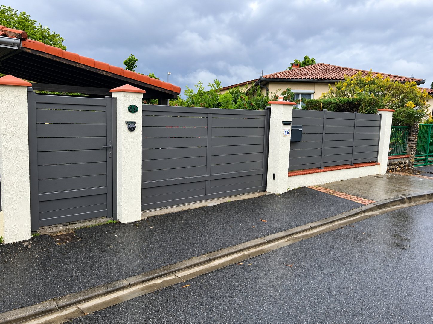Un portail coulissant, un portillon et une clôture en aluminium gris anthracite entre des piliers blancs à tuiles orange, devant une maison sous un ciel nuageux.