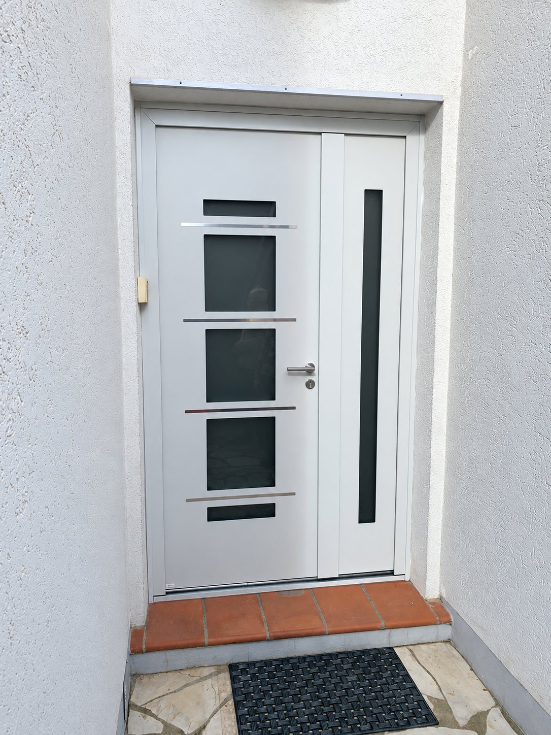 Porte d'entrée blanche moderne à quatre petits vitrages carrés et fin panneau latéral vitré, encadrée par un mur crépi, avec un seuil en carreaux de terre cuite.