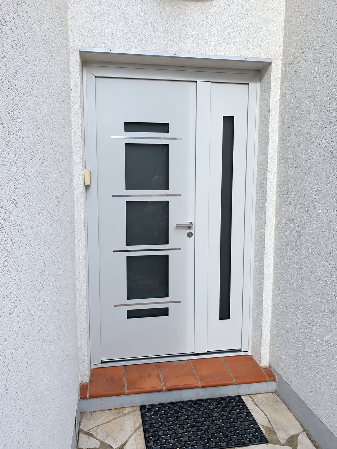 Porte d'entrée blanche moderne avec quatre petits vitrages sombres et un panneau latéral vitré, posée dans un renfoncement de façade crépie avec seuil en carreaux de terre cuite.