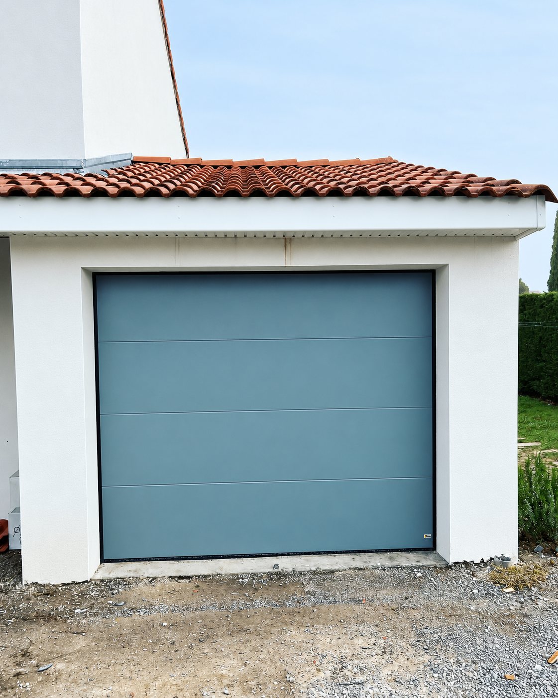 Une porte de garage sectionnelle gris-bleu installée dans un garage aux murs blancs surmonté d'un toit de tuiles rouges, devant une allée de gravier.