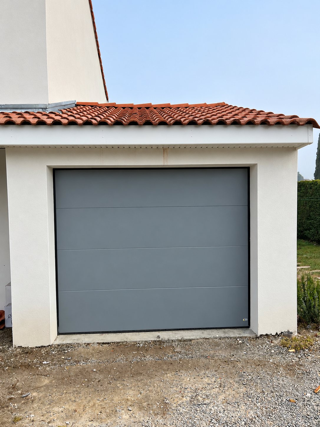 Une porte de garage sectionnelle grise posée dans l'encadrement d'un garage aux murs blancs et au toit de tuiles rouges.