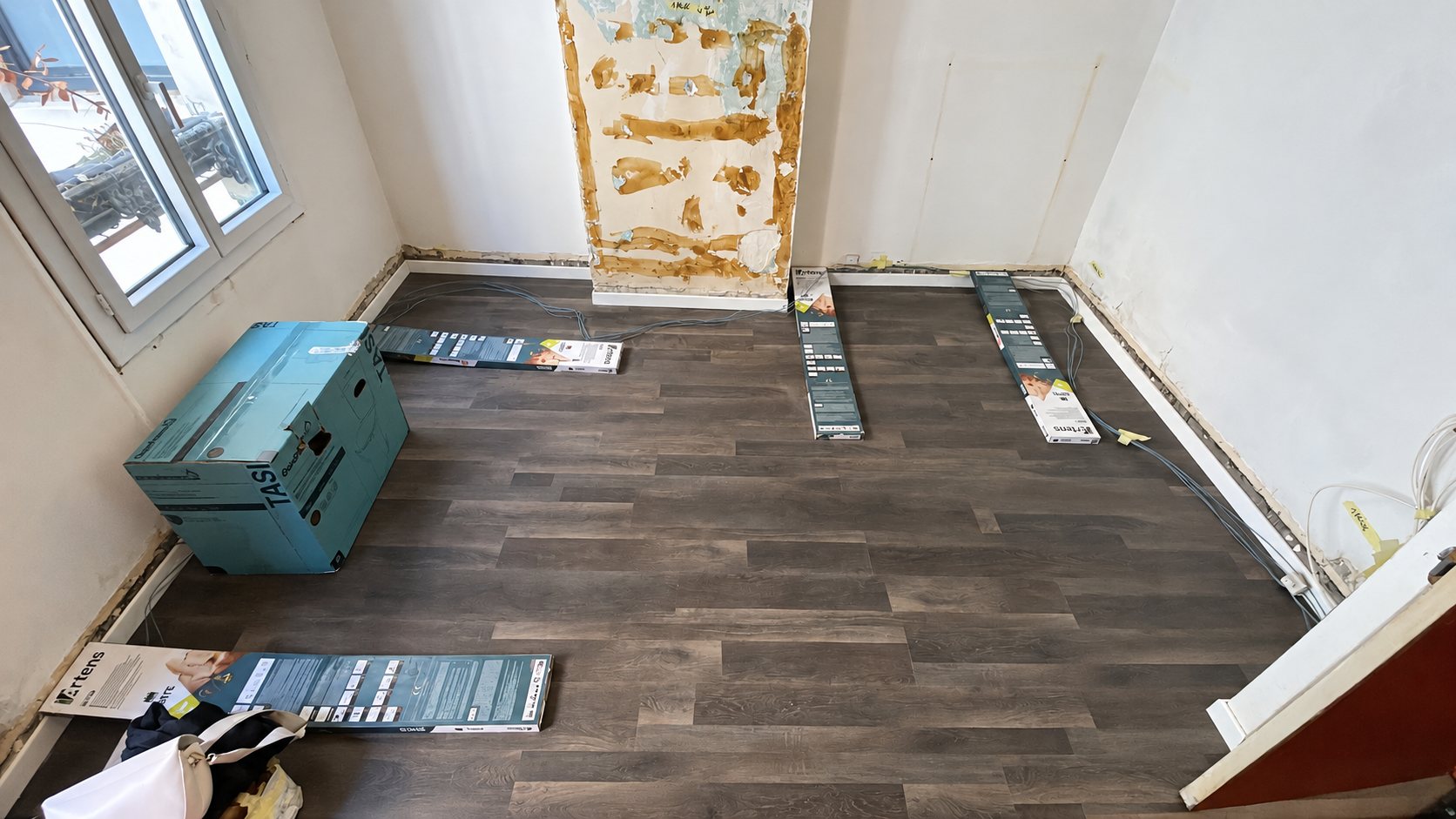 Pièce en rénovation dont le sol est recouvert de lames vinyle gris foncé, avec des paquets de lames posés dessus et un mur en cours de remise en état.