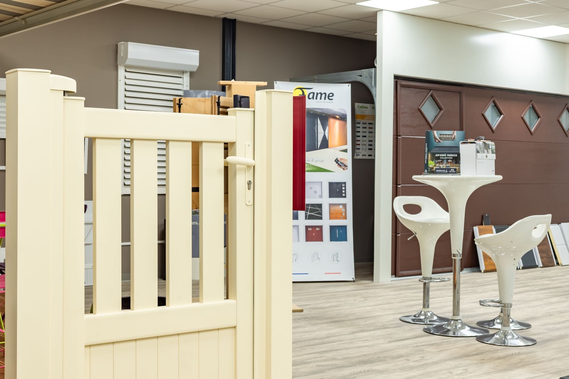 Intérieur d'un showroom présentant un portillon crème au premier plan, une porte de garage marron à losanges, une kakémono publicitaire et une table haute avec deux tabourets blancs.