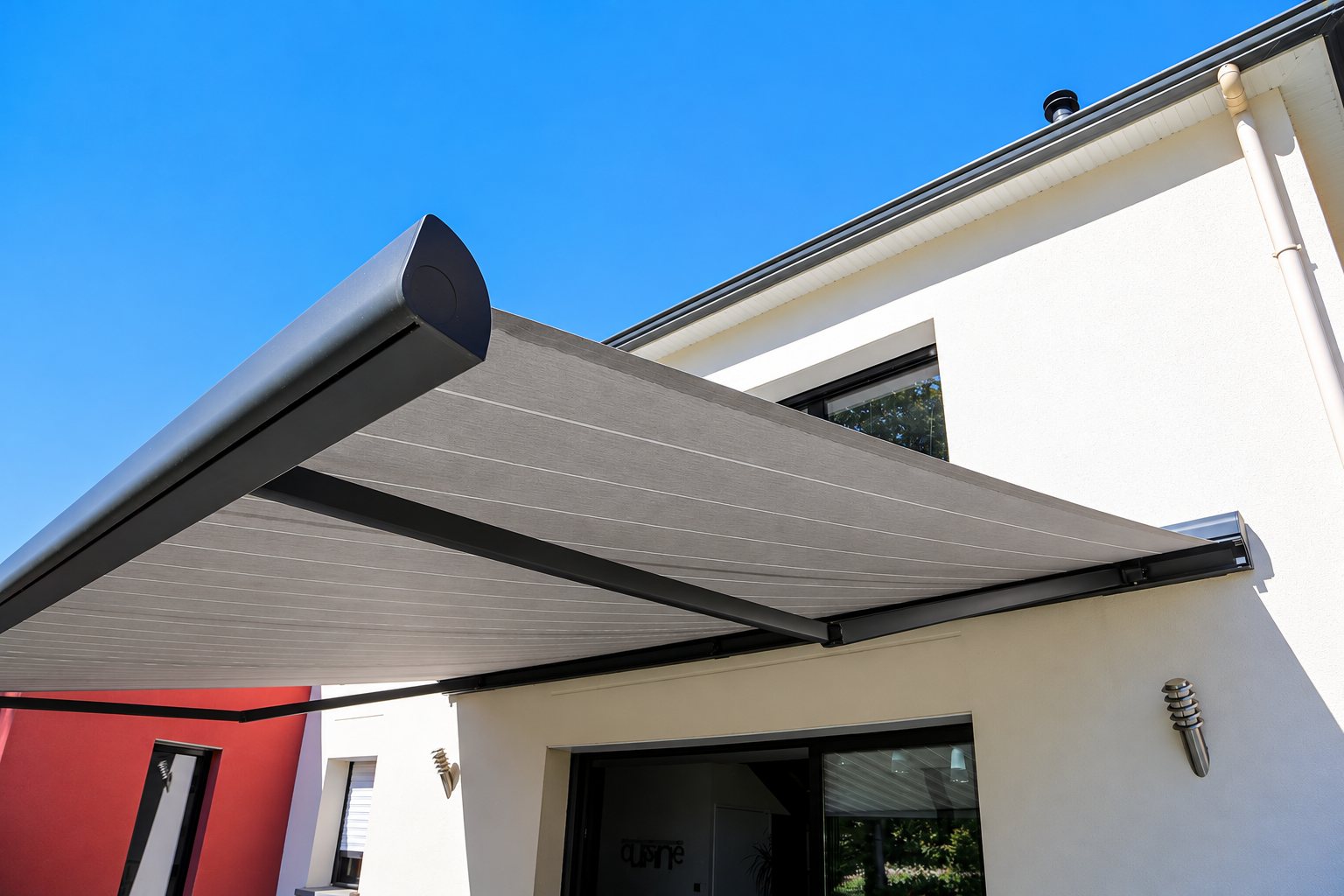 Un store banne gris anthracite déployé au-dessus d'une baie vitrée sur la façade blanche d'une maison contemporaine, sous un ciel bleu.
