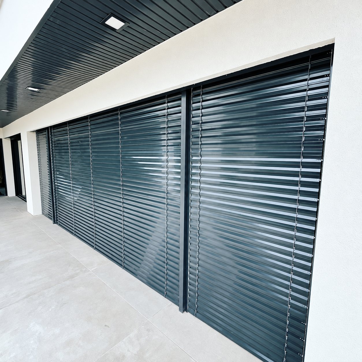 Grands stores à lamelles orientables de couleur gris-vert foncé installés sous un plafond de terrasse sur une façade blanche récente.
