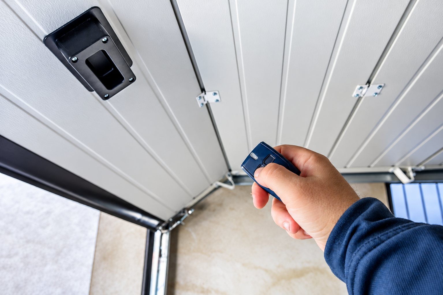 Une main tient une télécommande bleue pour actionner une porte de garage sectionnelle blanche vue de l'intérieur du garage.